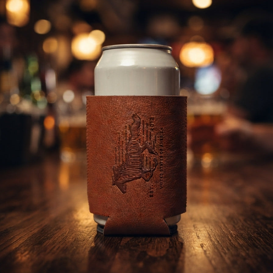 A brown leather can cooler sleeve with an embossed illustration of a pub interior, holding a white beverage can on a wooden bar table with a blurred bar background.